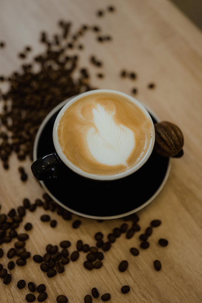 A warm cappuccino with elegant latte art served beside a macaron on a wooden table with scattered coffee beans.